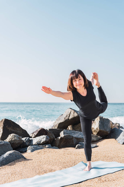 mujer feliz en ropa deportiva equilibrio en la pierna en Natarajasana. Ella está en una estera mientras hace yoga en la costa arenosa cerca del mar en un día soleado y mirando a la cámara - Foto, imagen