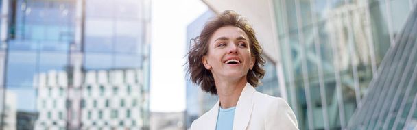 Усміхнена бізнес-леді в білому костюмі стоїть на хмарочосах міський пейзажний фон
 - Фото, зображення