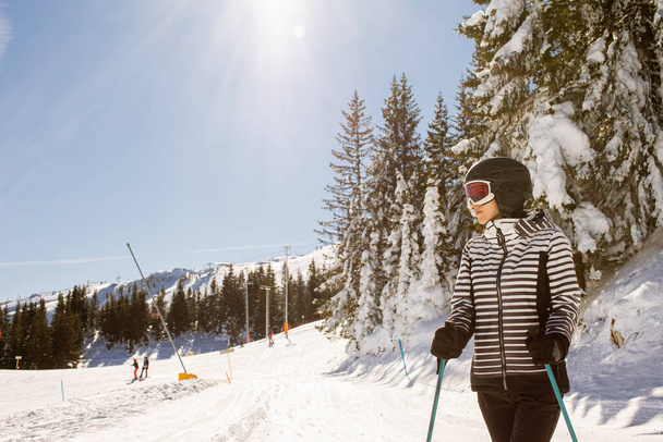 Νεαρή γυναίκα απολαμβάνει τη χειμωνιάτικη μέρα σκι στις χιονισμένες πλαγιές, περιτριγυρισμένη από ψηλά δέντρα και ντυμένη για κρύες θερμοκρασίες - Φωτογραφία, εικόνα