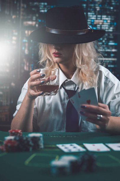 attraktives Mädchen in Hemd und Hut, das Whiskey trinkt und im Casino Pokerkarten betrachtet. Spielerin geht eine Wette ein. Poker im Casino - Foto, Bild