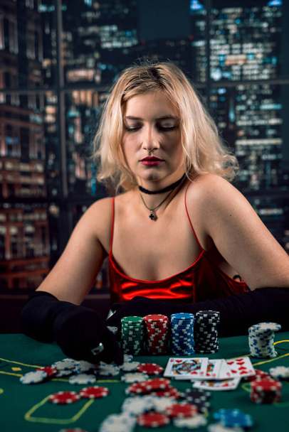 Kırmızı gece elbisesi giymiş duygusal genç bir bayan kumarhanede yeşil bir bezin üzerinde kağıt oynuyor. Poker oynayan bir kadın. kırmızı elbise - Fotoğraf, Görsel