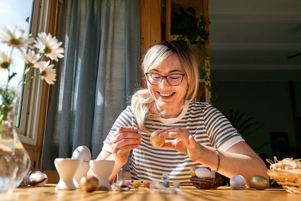 Улыбающаяся женщина готовит пасхальное украшение дома, рисует красочные пасхальные яйца и раскрашивает яичные чашки. - Фото, изображение