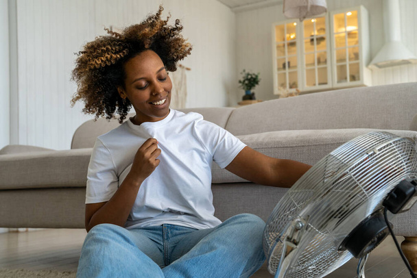 Satisfied joyful african american woman cools herself with electric fan rejoices in cool air hates stuffiness and heat. Pleased smiling female sit on floor holds regulates airflow from home ventilator - Zdjęcie, obraz