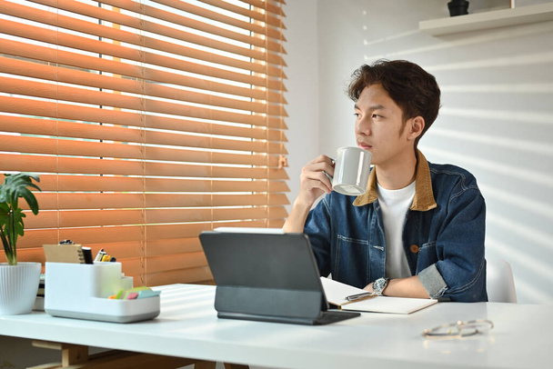 Προσεκτικός νεαρός Ασιάτης που πίνει καφέ και κοιτάζει από το παράθυρο, συλλογιζόμενος την ιδέα στο γραφείο του σπιτιού. - Φωτογραφία, εικόνα