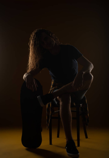 Jovem bonito macho sentado em uma cadeira e descansando a mão no skate no estúdio - Foto, Imagem
