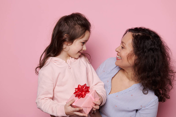 Anya és lánya jól érzik magukat együtt, mosolyognak, ölelkeznek, pózolnak egy aranyos ajándék elszigetelt rózsaszín háttér. International Womens Anyák Gyermeknapja koncepció. Család Emberek Emberi kapcsolatok - Fotó, kép