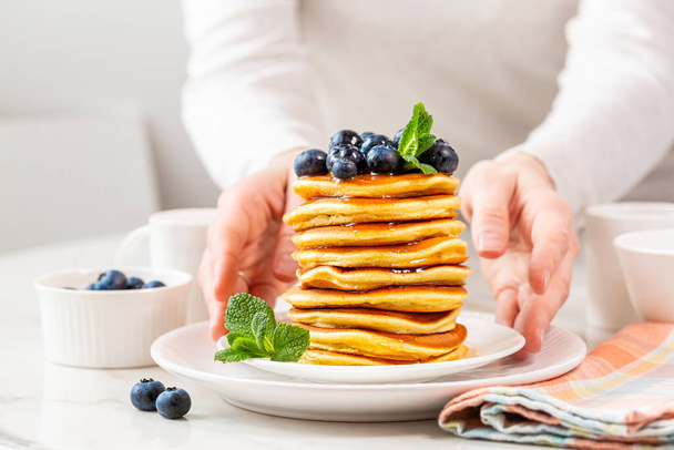 Morgens servieren Frauenhände hausgemachtes Frühstück, einen Stapel Pfannkuchen mit Blaubeeren und Honig oder Ahornsirup. Weißer Tisch Hintergrund. - Foto, Bild