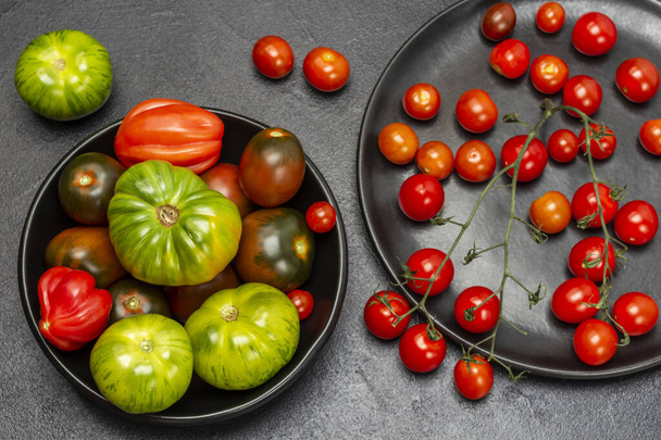 Red cherry tomatoes on a twig, green tomatoes in black plates. Flat lay. Black background. - Fotografie, Obrázek