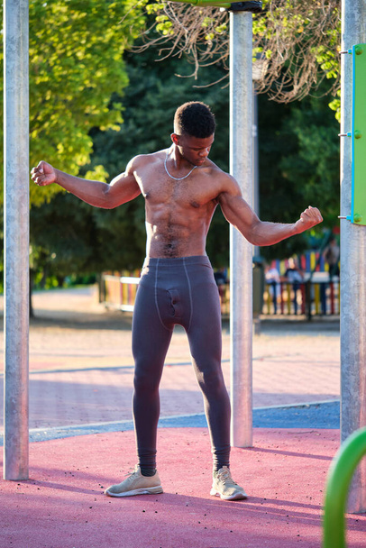 Nuori paidaton musta mies katselee lihaksiaan puistossa ulkona aurinkoisena päivänä. Kunto ja urheilu elämäntapa. - Valokuva, kuva