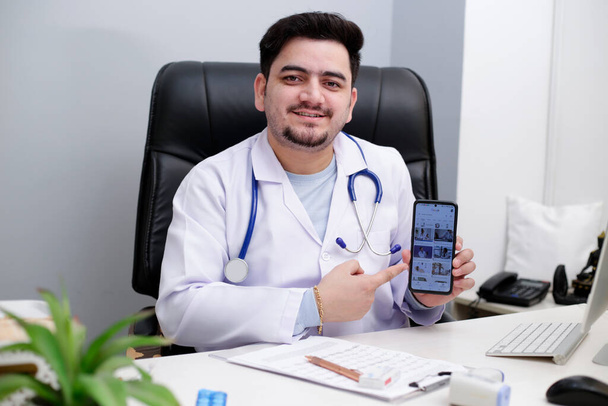 Ein junger Arzt sitzt auf dem Stuhl in der Klinik und zeigt etwas auf dem Handy. - Foto, Bild