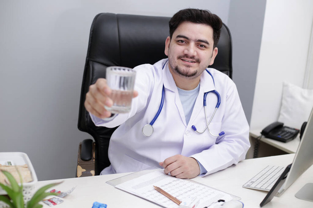 Um jovem médico está sentado na cadeira na clínica e segurando vidro de água na mão. - Foto, Imagem