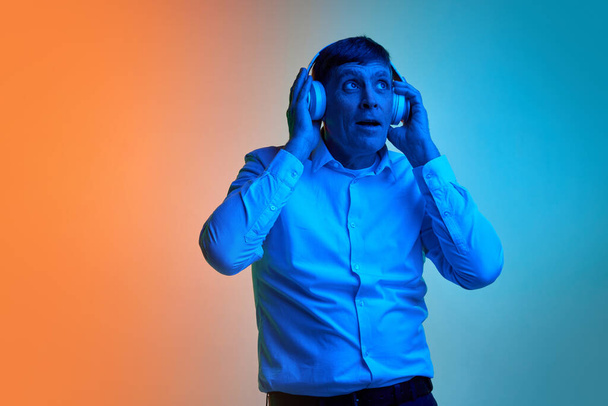 Portrait of mature, middle-aged man in white T-shirt listening to music in headphones posing over gradient blue orange background in neon light. Concept of emotions, lifestyle, facial expression, fun - Фото, зображення