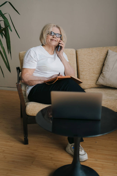 自宅でノートパソコンを使ってソファに座って仕事をしながら、カジュアルな服装で携帯電話で話をしたりメモを書いたりするシニア女性。遠隔地で働く実業家 - 写真・画像