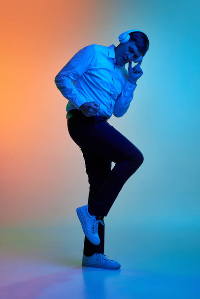 Full-length portrait of mature, middle-aged man listening to music in headphones and dancing over gradient blue orange background in neon light. Concept of emotions, lifestyle, facial expression, fun - Foto, afbeelding