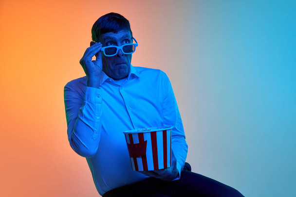 Ritratto di uomo maturo di mezza età in posa in occhiali 3D e popcorn su sfondo blu arancio sfumato in luce al neon. Un film impressionante. Concetto di emozioni, stile di vita, espressione facciale, divertimento. Pubblicità - Foto, immagini