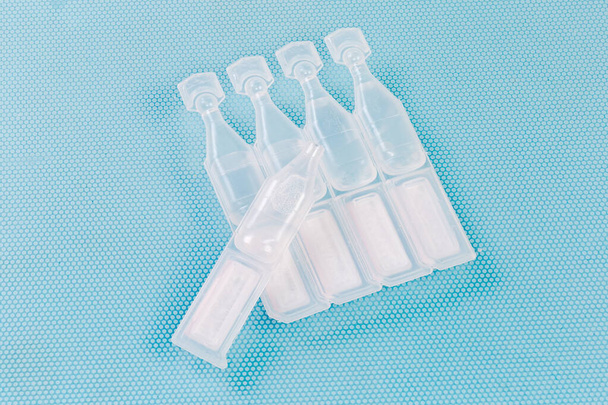 Gotas para los ojos en pequeñas ampollas de plástico desechables conectadas en bloque y una ampolla abierta por separado, primer plano sobre una superficie azul - Foto, Imagen