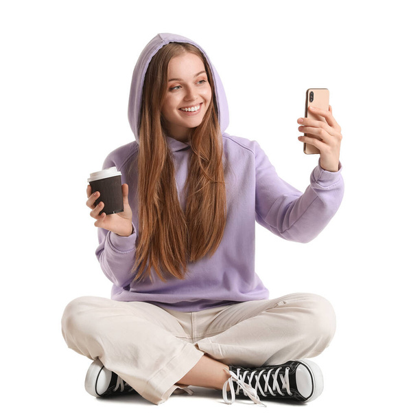 Hermosa chica con taza de café y teléfono móvil tomando selfie sobre fondo blanco - Foto, imagen