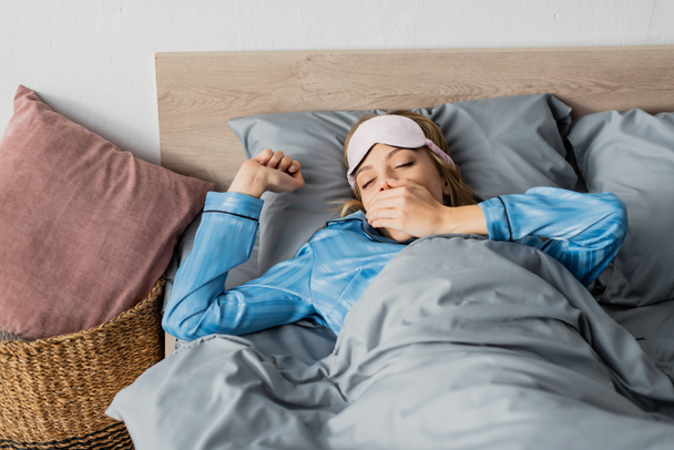 сонна жінка в шовковій піжамі і нічній масці позіхання і прикриття рота, лежачи в ліжку
  - Фото, зображення