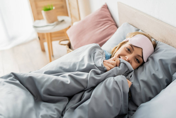 Ευτυχισμένη νεαρή γυναίκα με μάσκα ύπνου ξαπλωμένη κάτω από κουβέρτα σε άνετο κρεβάτι  - Φωτογραφία, εικόνα