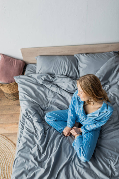 Draufsicht auf eine barfüßige und fröhliche Frau im blauen Pyjama, die auf dem Bett sitzt  - Foto, Bild