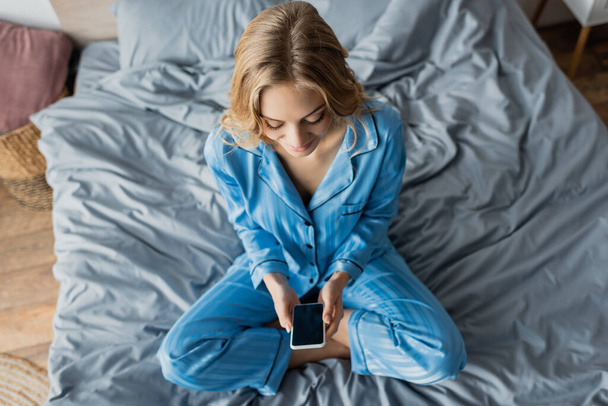vue du dessus de la femme joyeuse en pyjama bleu en utilisant un smartphone avec écran vide tout en étant assis sur le lit  - Photo, image