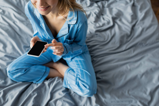 vista recortada de la mujer sonriente en pijama azul usando un teléfono inteligente con pantalla en blanco mientras está sentada en la cama  - Foto, Imagen