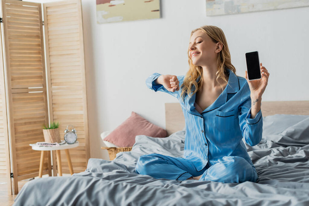 pleased woman in blue pajama holding smartphone with blank screen  - Zdjęcie, obraz