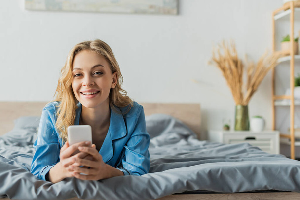 joyful young woman messaging on smartphone while lying in blue pajama on bed  - Фото, зображення