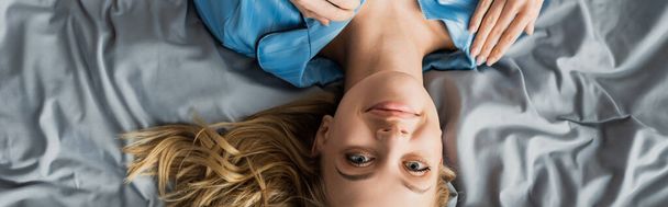 vista superior de la mujer despreocupada en pijama de seda azul acostada en la cama y mirando a la cámara, pancarta  - Foto, Imagen