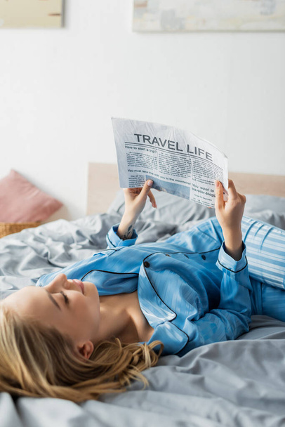 tattooed young woman smiling while reading travel newspaper in bed  - Photo, Image