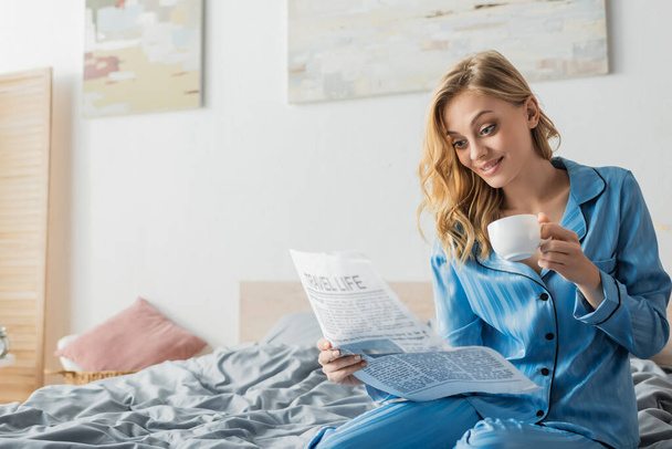amazed young woman reading travel life newspaper and holding cup of coffee in bed  - Foto, Imagem