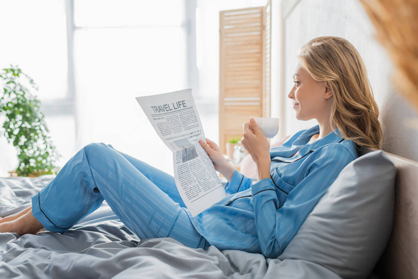 vista lateral de la joven despreocupada leyendo el periódico de viaje y sosteniendo la taza de café en la cama  - Foto, imagen