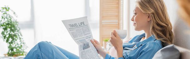 side view of carefree young woman reading travel life newspaper and holding cup of coffee in bedroom, banner  - Foto, imagen