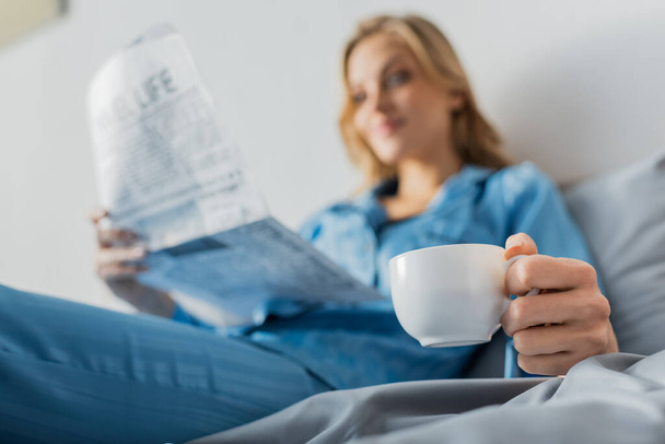 Bulanık arka planda gazetesi olan bir kadının elinde bir fincan kahve.  - Fotoğraf, Görsel