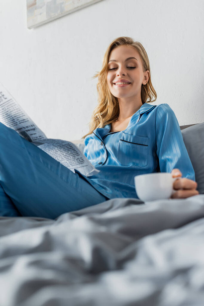 šťastná mladá žena drží noviny a při pohledu na šálek kávy, zatímco odpočívá v posteli  - Fotografie, Obrázek