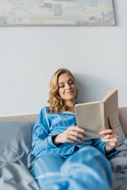 mujer joven y complacida en azul de seda camisón libro de lectura en el dormitorio  - Foto, Imagen