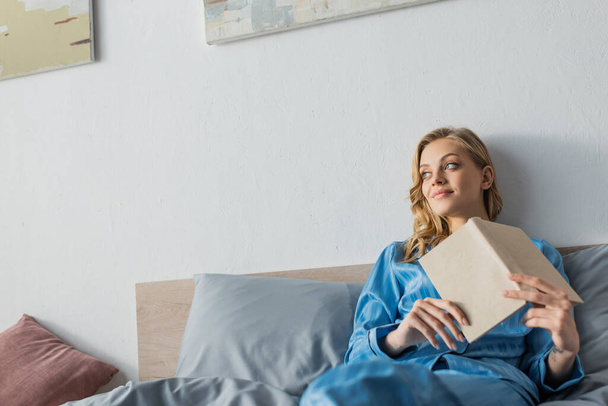 青い絹のナイトウェアを着た若い女性が寝室で本を持っています  - 写真・画像