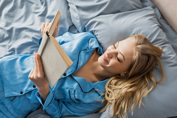 top view of pleased young woman in blue silk nightwear reading book on bed  - Foto, imagen
