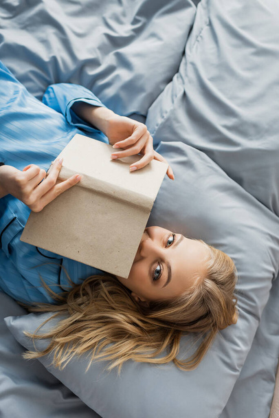 vista superior de la joven complacida en la cara de la cubierta de la ropa de dormir de seda azul con libro en la cama  - Foto, imagen
