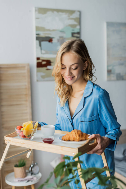 femme heureuse en pyjama bleu regardant plateau avec petit déjeuner dans la chambre  - Photo, image
