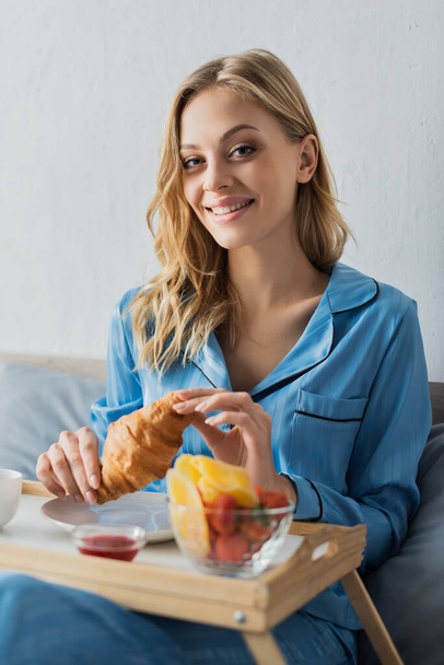 pretty woman in blue pajama holding fresh croissant near tray while having breakfast in bed  - Fotoğraf, Görsel