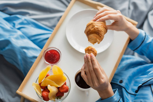 vista superior de la mujer recortada sosteniendo croissant fresco cerca de bandeja mientras desayuna en la cama  - Foto, imagen