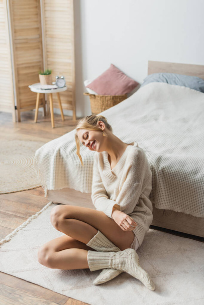 young blonde woman in knitted sweater and socks sitting on carpet in modern bedroom  - Fotografie, Obrázek