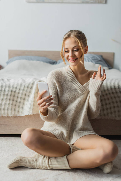 cheerful young woman in sweater having video call on smartphone in modern bedroom  - Photo, Image