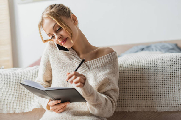 mulher bonita em suéter falando no smartphone e tomar notas no notebook  - Foto, Imagem
