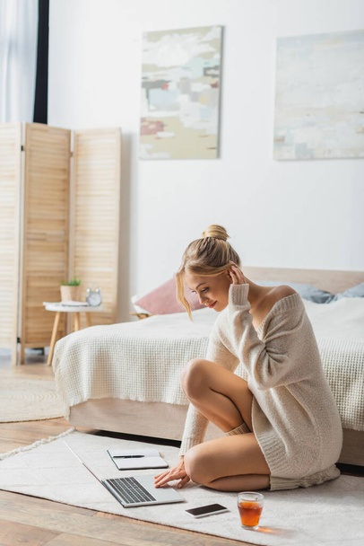 charming woman in sweater using laptop near mobile phone and cup of tea on carpet in bedroom  - Foto, Bild