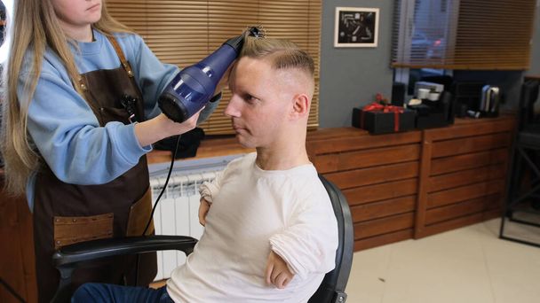 uma menina cabeleireiro em um salão de beleza faz estilo de cabelo para um homem com deficiência - Foto, Imagem