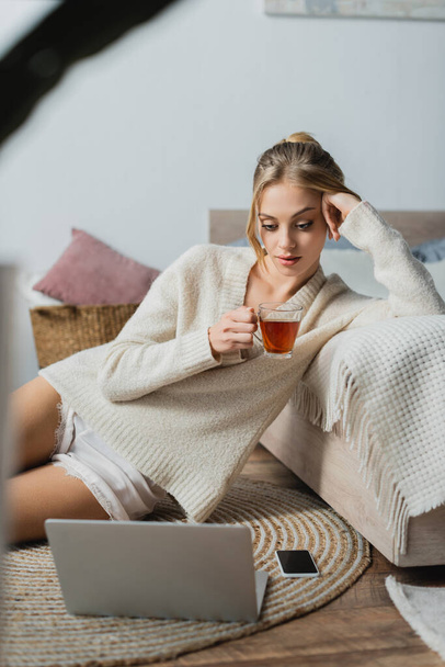 young blonde woman in sweater holding glass cup with tea and looking at laptop in bedroom  - Zdjęcie, obraz