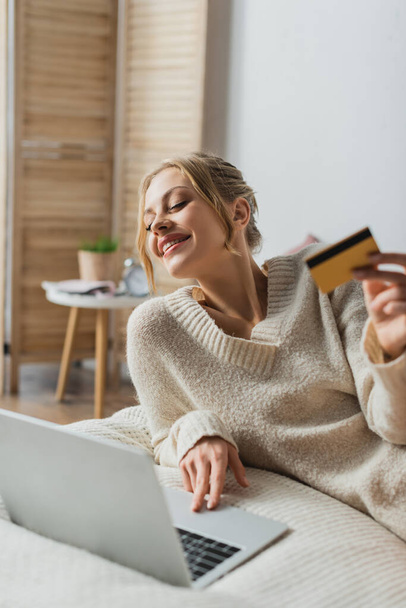 ευχαριστημένη γυναίκα κρατώντας πιστωτική κάρτα και χρησιμοποιώντας φορητό υπολογιστή, ενώ κάνει online ψώνια στην κρεβατοκάμαρα  - Φωτογραφία, εικόνα