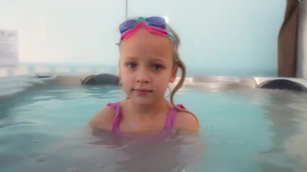 Пятилетняя девочка с очками для плавания на лбу сидит в джакузи на открытом воздухе и смотрит в камеру, а потом чихает - Кадры, видео